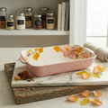Decorative pink baking dish with floral design on a marble surface, surrounded by spices and books.